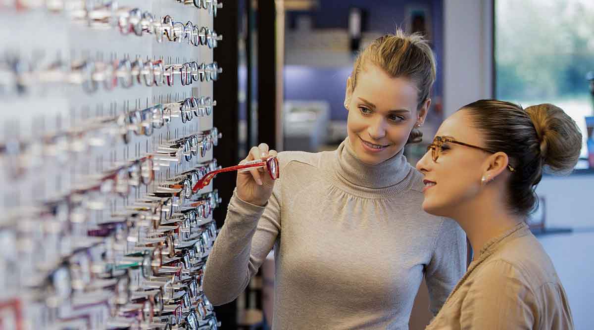 Duas mulheres jovens olhando mostruário de óculos de grau numa loja de uma franquia de ótica.