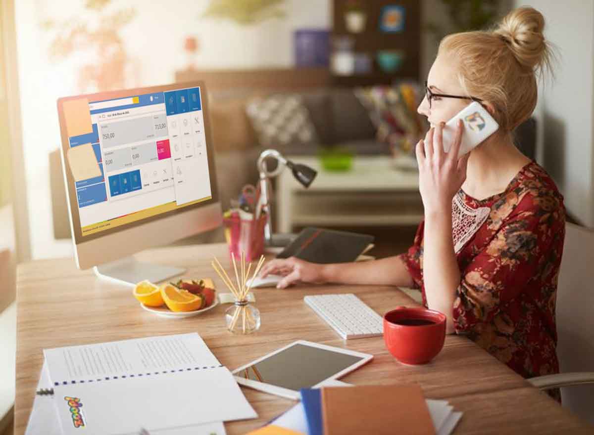 Uma mulher loira, jovem e usando óculos fala ao telefone enquanto olha para a tela do seu computador para trabalhar como freelancer.