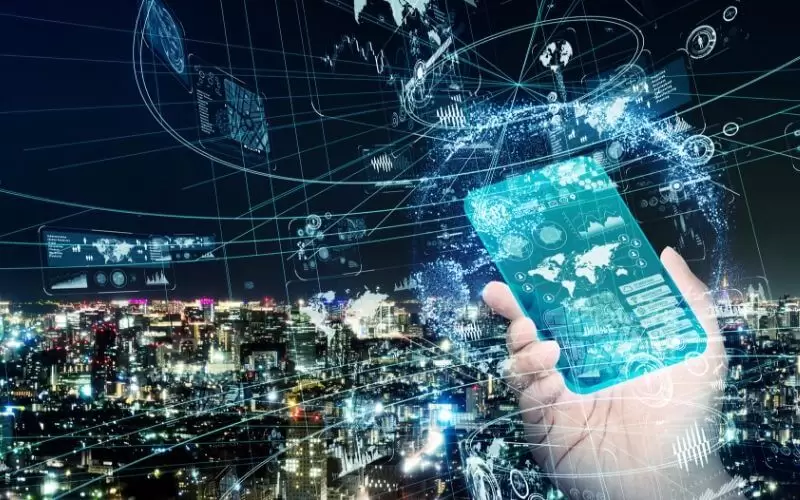 Imagem mostra uma mão humana segurando um celular tendo ao fundo uma cidade conectada